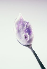Łyżka zawierająca kremowy jasnofioletowy substancję z małymi fioletowymi kwiatkami i kryształkami, ustawiona na tle w delikatnym, gradientowym kolorze.