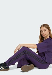 Purple casual outfit featuring a loose t-shirt and track pants with black side stripes. Black sneakers with white accents. Smooth fabric texture.