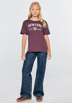 Blondynka stoi ubrana w fioletowy t-shirt z napisem New York, luźne niebieskie dżinsy i fioletowe buty na jednolitym jasnym tle.