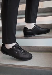 Černé tenisky s texturovanou kůží a semišovými akcenty, s kulatými tkaničkami, nízkým profilem a značeným logem na boku.