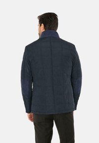 Giacca trapuntata blu navy con colletto alto, toppe sui gomiti a contrasto e superficie liscia, caratterizzata da un taglio dritto e dettagli delle cuciture sul retro.