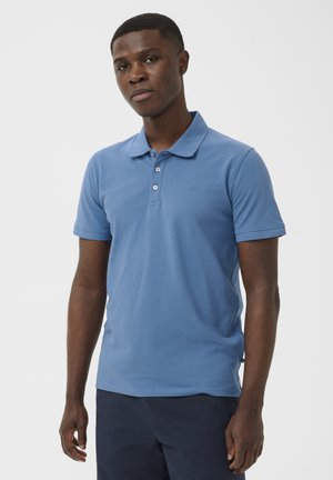 Jeune homme portant un polo bleu clair à manches courtes et un pantalon foncé, debout devant un fond blanc uni.