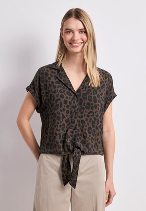 Smilende kvinde med glat blondt hår iført en kortærmet brun leopardprint bluse, bundet i taljen, og beige bukser mod hvid baggrund.