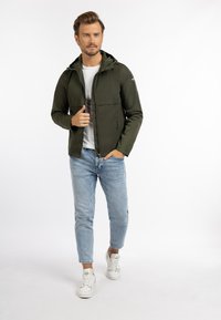 Olivengrøn letvægtsjakke med frontlynlås, hoodie og brystlomme, båret over en hvid t-shirt og parret med lyseblå jeans og sneakers.