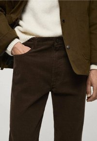 Bruine corduroy broek met een textuurafwerking, voorzien van een voorzak en knoopsluiting, gecombineerd met een witte trui en een donkere jas.