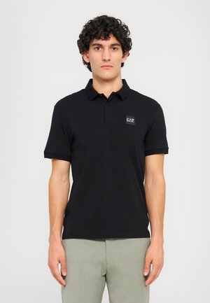 Black polo shirt with short sleeves and a collared neck. Features a small EA7 logo patch on the chest. Cotton material, smooth texture.