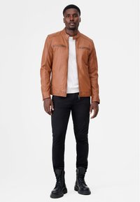Brun læderjakke med opretstående krave, lynlåse foran og en tekstureret overflade. Bæres over en hvid striktrøje og parret med sorte jeans og støvler.