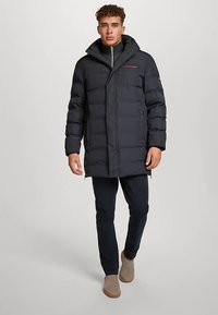 Mørk dunjakke med hette, med glidelås foran, elastiske mansjetter og en logo på brystet. Brukt over mørke bukser og beige støvler.