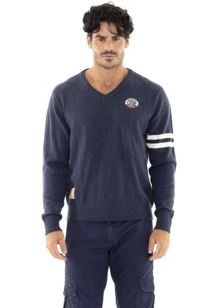 Un homme porte un pull bleu marine à col en V, orné de rayures blanches sur les manches et d’un logo sur la poitrine, avec un pantalon cargo bleu marine, sur fond blanc.