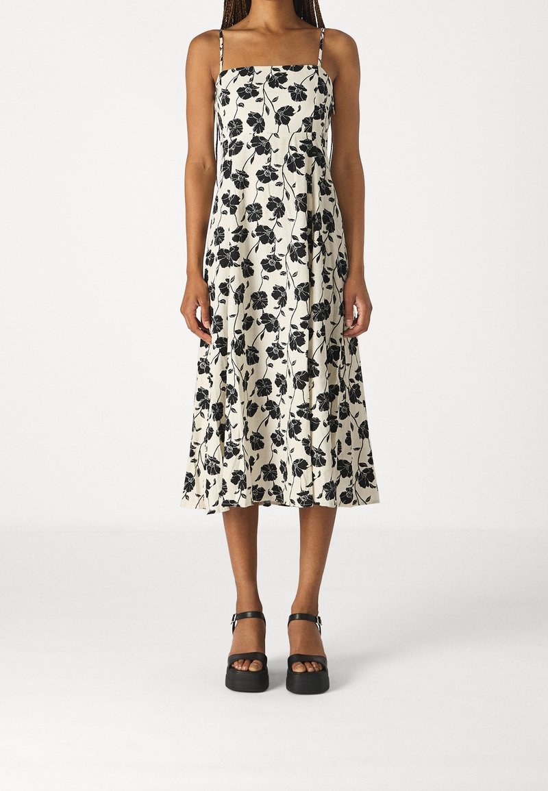 Femme portant une robe midi blanche à motif floral noir avec fines bretelles, associée à des sandales à plateforme noires.