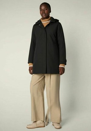 Donna con cappotto nero con cappuccio, maglione dolcevita a righe marroni, pantaloni beige a gamba larga e scarpe beige, in piedi contro uno sfondo semplice.