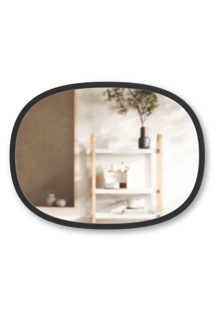 Specchio ovale con una cornice nera, che riflette uno scaffale sfocato con struttura in legno, scaffali bianchi e vari piccoli oggetti decorativi.