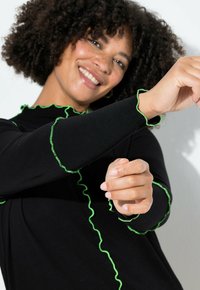 Zwarte top met lange mouwen, voorzien van groene ruches langs de halslijn en manchetten, gemaakt van een gladde, rekbare stof. Handen speels gepositioneerd.