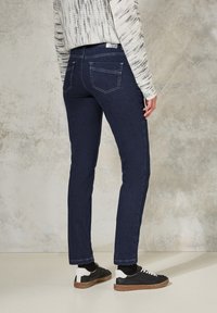 Jean bleu foncé avec une coupe slim, présentant une taille intermédiaire et deux poches arrière. Associés à des baskets noires avec des accents blancs.