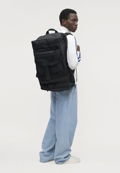 Un homme en chemise blanche et en jean ample porte un grand sac à dos noir et regarde par-dessus son épaule devant un fond de studio uni.