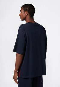 Młody mężczyzna z krótkimi kręconymi włosami, ubrany w luźną granatową koszulkę i pasujące spodnie, stojący bokiem na jednolitym tle.