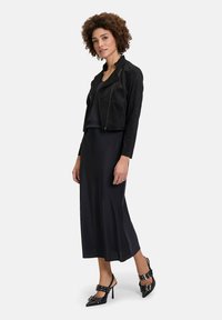 Schwarze Wildleder-Cropped-Jacke mit asymmetrischem Reißverschluss, kombiniert mit einem langen schwarzen Satinkleid und schwarzen Stiletto-Heels mit Schnallelementen.