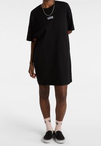 Czarna sukienka T-shirt o luźnym kroju, z białym logo Vans na klatce piersiowej, zestawiona z różowymi skarpetkami i czarnymi butami slip-on.