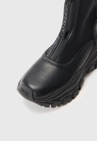 Czarne wodoodporne buty z zaokrąglonym czubkiem, fakturowaną cholewką i solidną podeszwą. Wyposażone w zamek błyskawiczny i detal z logo po boku.