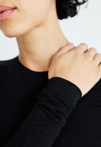 Close-up van een persoon in een zwart shirt met lange mouwen, met één hand op de tegenovergestelde schouder, tegen een witte achtergrond.