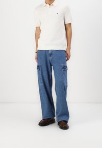 Osoba v bílém polo tričku s krátkým rukávem, volných modrých cargo džínách s kapsami a tmavě hnědých kožených botách stojící na bílém pozadí.