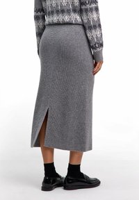 Person, die einen grauen gerippten Midi-Rock mit einem Schlitz hinten, schwarze Socken, schwarze Loafers und einen gemusterten grauen Pullover trägt, steht auf weißem Boden.