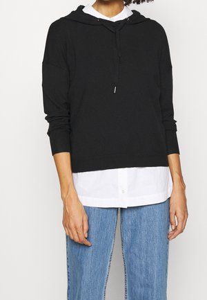 Sweatshirt à capuche noire avec des poignets côtelés, ornée d'un col à cordons, superposée à une chemise blanche à boutons et accompagnée d'un jean bleu.