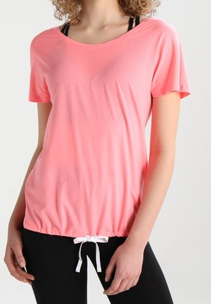 Kobieta ubrana w luźną różową bluzkę z krótkim rękawem ze białym sznurkiem na dole oraz czarne legginsy na neutralnym tle.