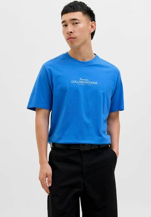 Blaues Baumwoll-T-Shirt mit kurzen Ärmeln und Rundhalsausschnitt, mit weißem Text "Premium COLLABORATIONS FOUNDED IN 1990" auf der Brust. Schwarze Hose.