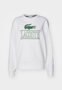 Felpa bianca con logo di coccodrillo verde e testo "LACOSTE". Maniche lunghe, scollo rotondo e texture morbida e liscia.