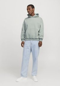 Halványzöld kapucnis pulóver kenguruzsebbel, világoskék, laza fazonú farmerrel és fehér sportcipővel viselve. Sima textúra, hétköznapi dizájn.