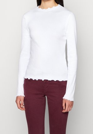 Personne portant un haut blanc à manches longues en tissu côtelé avec des bords festonnés et un pantalon rouge foncé, debout devant un fond clair uni.