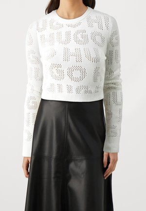Pull en maille blanc à manches longues avec lettrage perforé "HUGO" porté avec une jupe en cuir noire taille haute, fond clair, visage non visible.