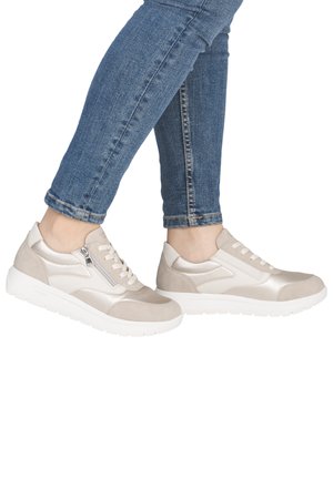 Person wearing beige and white casual sneakers with side zippers and blue jeans, feet crossed at the ankles on a white background.