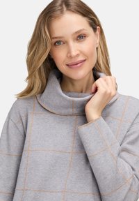 Kobieta z blond włosami, ubrana w szary sweter z golfem w jasnobrązowe kratki, delikatnie trzymająca kołnierz, uśmiechająca się łagodnie.