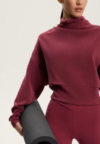 Hanorac bordeaux cu guler înalt, mâneci largi și talie ajustată, completat cu colanți asortați, ținând un covoraș de yoga texturat negru.
