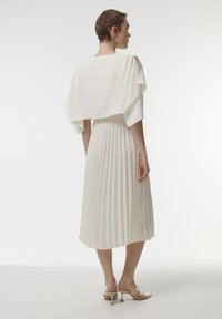 Witte geplooide jurk met een vloeiende cape mouwontwerp, gemaakt van een lichtgewicht stof. Heeft een subtiele textuur en een jurkzoom tot de knie.