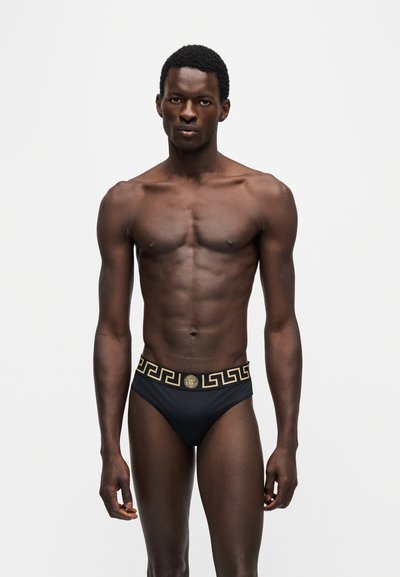 Homme debout face à l'avant portant un slip noir avec motif grec doré et ceinture ornée de l'emblème Méduse, sur fond uni clair.