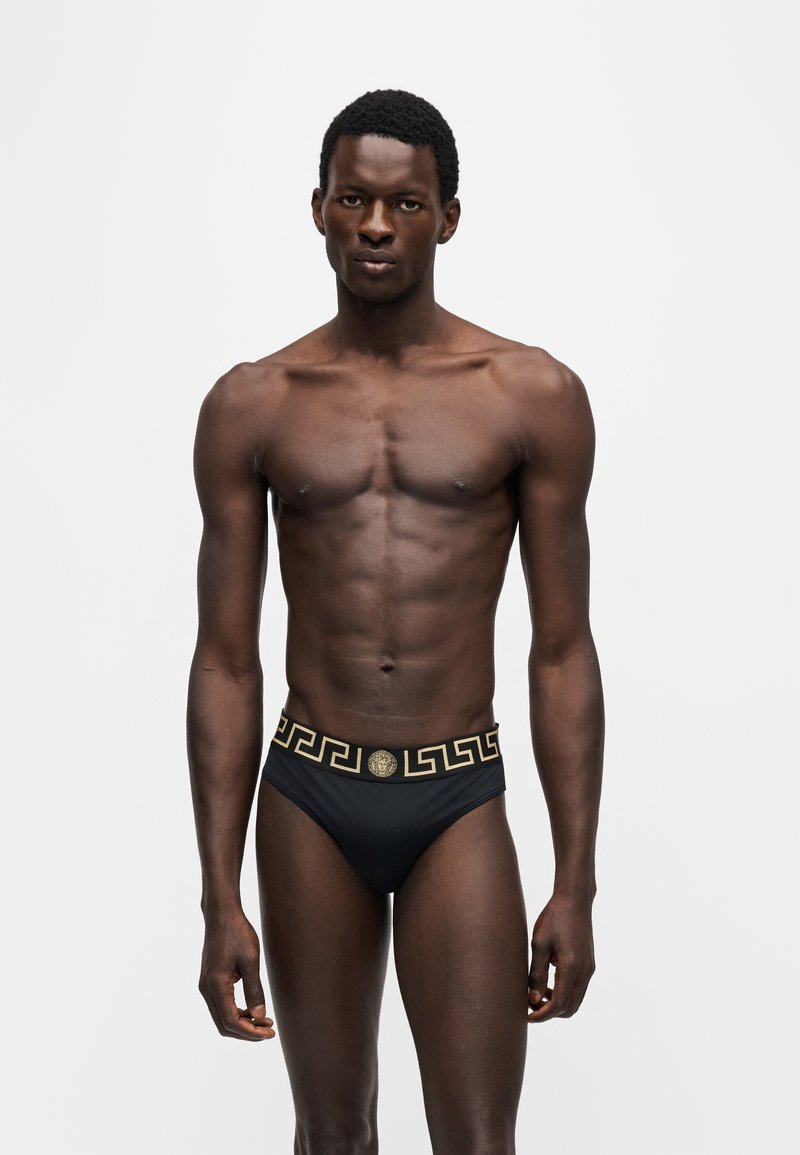 Homme debout face à l'avant portant un slip noir avec motif grec doré et ceinture ornée de l'emblème Méduse, sur fond uni clair.