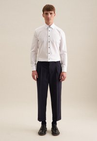 Camicia bianca con bottoni neri, abbinata a pantaloni plissettati blu scuro. Scarpe eleganti nere e calzini blu. Sfondo semplice.