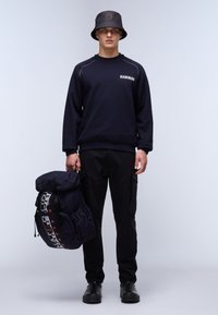 Felpa navy con cuciture a contrasto, pantaloni cargo neri e un cappello a secchiello scuro. Tiene uno zaino scuro e textured con accenti di logo.