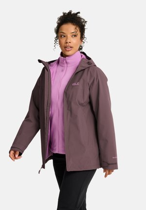 Vrouw draagt een mauve waterdichte jas over een lichtpaarse fleece met rits, staat en kijkt iets opzij op een witte achtergrond.