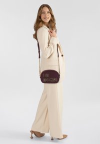 Borsa a tracolla bordeaux con il logo "LOVE MOSCHINO" dorato, texture liscia e dettagli con tracolla a catena, indossata con un completo beige su uno sfondo neutro.