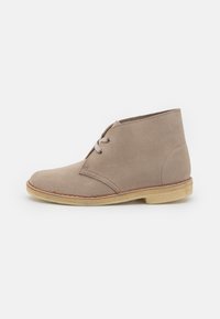 Beige suede ankle boots with a rounded toe, two eyelets, and light-colored rubber sole. Features stitched detailing along the edge.