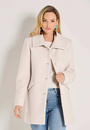 Femme portant un manteau en laine beige clair avec un grand col, des boucles d’oreilles colorées surdimensionnées, un pull blanc et un jean bleu sur fond uni.