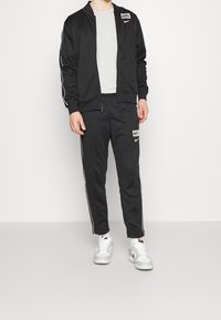 Černý Nike teplákový komplet vyrobený z hladkého materiálu, s bílými bočními pruhy, zateplenou bundou na zip, logem na hrudi a nohavicích, doplněný bílými teniskami.