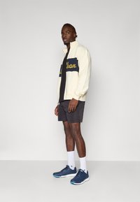 Cremefarvet zip-jakke med marineblå panel og gul tekst, sort t-shirt, mørke shorts, hvide sokker og blå sportssko. Enkel design.