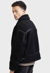 Schwarze Fake-Shearling-Jacke mit Lederakzenten, einem Kragen und einem kastigen Schnitt. Verfügt über eine dicke Textur und ein mittel-langes Design.