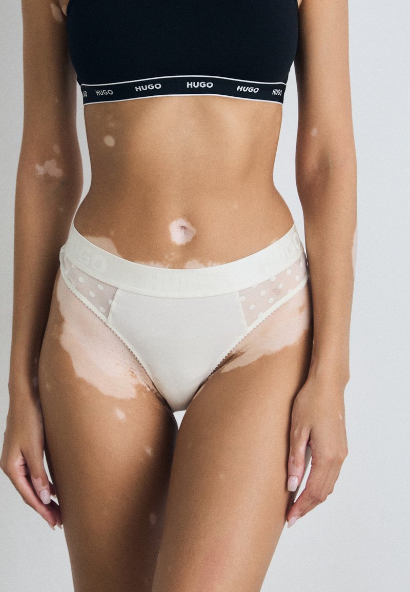 Culotte de bikini blanche avec un panneau latéral en mesh à pois transparent et une taille douce avec logo. Texture lisse, design taille mi-haute.