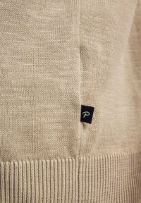 Beige Strickpullover mit geripptem Saum und einem kleinen Marineblau-Label mit einem weißen Logo, das an der Seite aufgestickt ist. Glatte Textur, leichtes Material.